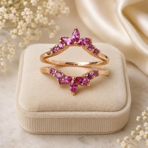 May include: A rose gold ring guard set featuring two curved bands adorned with vibrant pink marquise and round cut stones. The design creates a symmetrical, pointed crown-like shape, with the stones set along the top edge of each band.