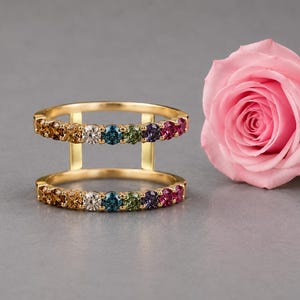 May include: A gold double-band ring featuring two parallel rows of multi-colored gemstones, including shades of brown, white, blue, green, purple, and pink, set in a prong setting. The ring is positioned next to a single light pink rose.