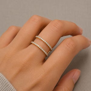 May include: A gold double-band ring featuring two parallel rows of small, clear, round-cut stones set along the entire length of each band. The two bands are connected by a small vertical bar on one side, creating an open, layered appearance.
