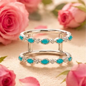 May include: A silver ring with two bands, each adorned with oval turquoise stones and small clear gems. The ring is displayed with pink roses and petals, creating a romantic and elegant aesthetic.