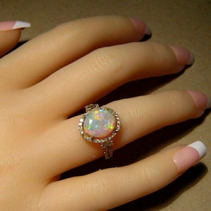 May include: A gold ring with a large, oval opal gemstone, surrounded by a halo of small, clear gemstones. The opal displays a play of color with flashes of blue, green, and yellow. The ring is on a hand with a French manicure.