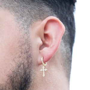 May include: Gold hoop earring with a dangling cross charm, both adorned with clear cubic zirconia stones.