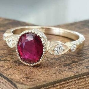Puede incluir: Un anillo de oro con un gran rubí ovalado de color rojo intenso. El rubí está engastado en un bisel texturizado. Pequeñas piedras preciosas transparentes están engastadas a ambos lados del rubí, añadiendo brillo.