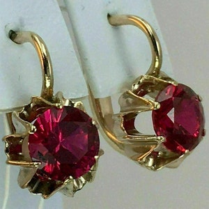 May include: A pair of gold earrings with red gemstone accents. The earrings feature a round, faceted gemstone set in a gold setting with a delicate, ornate design.