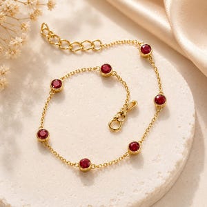 May include: A gold chain bracelet featuring six evenly spaced, round, deep red faceted stones set in gold bezels. The bracelet includes a delicate link chain, a secure clasp mechanism, and an adjustable extension chain with several circular links.