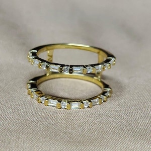 Op de afbeelding: Twee goudkleurige ringen boven elkaar. Elke ring is versierd met een reeks kleine, rechthoekige en ronde, heldere edelstenen. De ringen zijn geplaatst tegen een neutrale, beige achtergrond.