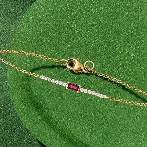 May include: A delicate gold bracelet with a small, rectangular ruby center stone. The bracelet features a chain design with small, sparkling white stones on either side of the ruby. The clasp is gold and the background is a solid green.