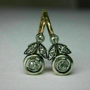 May include: A pair of silver dangle earrings with a round diamond in the center of each earring. The earrings are accented with smaller diamonds and have a gold hook.