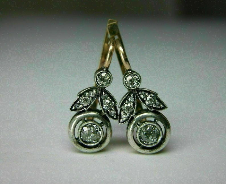 May include: A pair of silver dangle earrings with a round diamond in the center of each earring. The earrings are accented with smaller diamonds and have a gold hook.