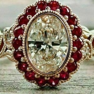 May include: An ornate gold ring featuring a large, oval-cut diamond center stone surrounded by a halo of small, round red gemstones. The ring's band has intricate filigree detailing.