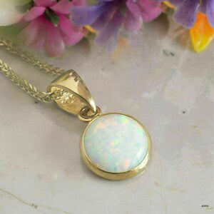 Puede incluir: Un collar con colgante de oro con una piedra preciosa de ópalo redonda e iridiscente. El ópalo está engastado en un bisel de oro y cuelga de una fina cadena de oro. El fondo incluye flores coloridas.