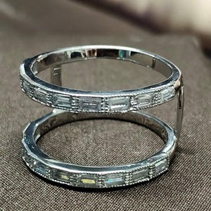 Puede incluir: Un anillo plateado con dos bandas paralelas unidas por barras verticales. Cada banda está adornada con piedras rectangulares y transparentes. El anillo, con su superficie reflectante, se destaca sobre un fondo marrón.