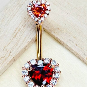 May include: Gold belly button ring with two heart-shaped settings. The top setting has an orange gemstone and the bottom setting has a red gemstone. Both settings are surrounded by clear cubic zirconia stones.