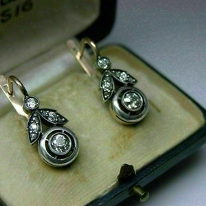 May include: A pair of silver and gold dangle earrings with a round diamond in the center and smaller diamonds around the edges. The earrings are in a small box.
