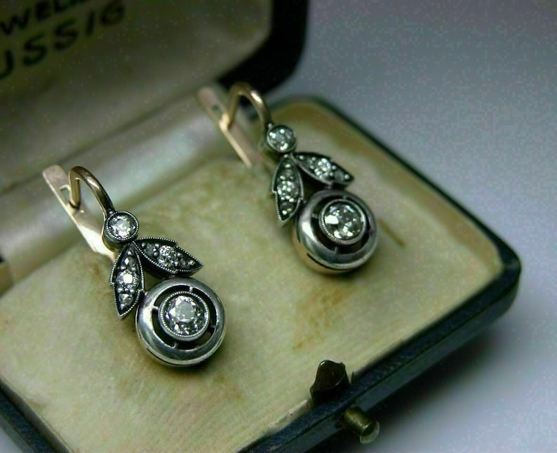 May include: A pair of silver and gold dangle earrings with a round diamond in the center and smaller diamonds around the edges. The earrings are in a small box.