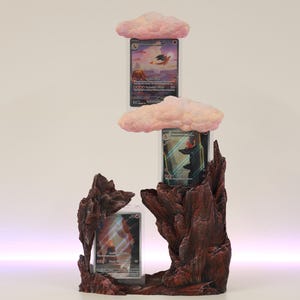 May include: A 3D printed display stand for trading cards, featuring a brown, rocky base with pink clouds and card slots. The stand is designed to hold three cards, with the top slot featuring a pink cloud and the bottom two slots featuring a brown rocky base.