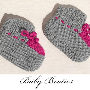 Può includere: Un paio di scarpine da neonato grigie lavorate a maglia con dettagli rosa acceso. Le scarpine hanno un polsino a coste e una sezione rosa sul davanti con filo rosa decorativo. Il testo "Baby Booties" è scritto sotto.