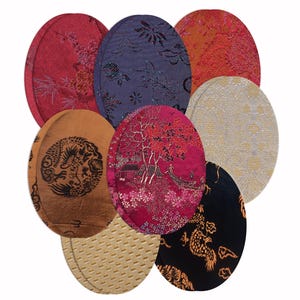 Könnte beinhalten: Eine Sammlung von acht ovalen Stoffuntersetzern mit verschiedenen Mustern und Farben, darunter Rot, Violett, Orange, Schwarz und Creme. Die Designs zeigen komplizierte Blumenmotive, Drachenillustrationen und strukturierte geometrische Muster.