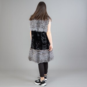 A female model is posing in front of a gray panel. She is wearing a vest made from real fox and mink fur in black and silver colors.