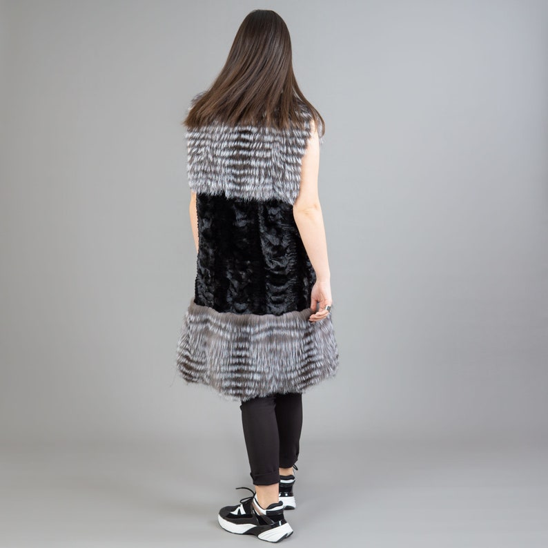 A female model is posing in front of a gray panel. She is wearing a vest made from real fox and mink fur in black and silver colors.