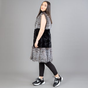 A female model is posing in front of a gray panel. She is wearing a vest made from real fox and mink fur in black and silver colors.
