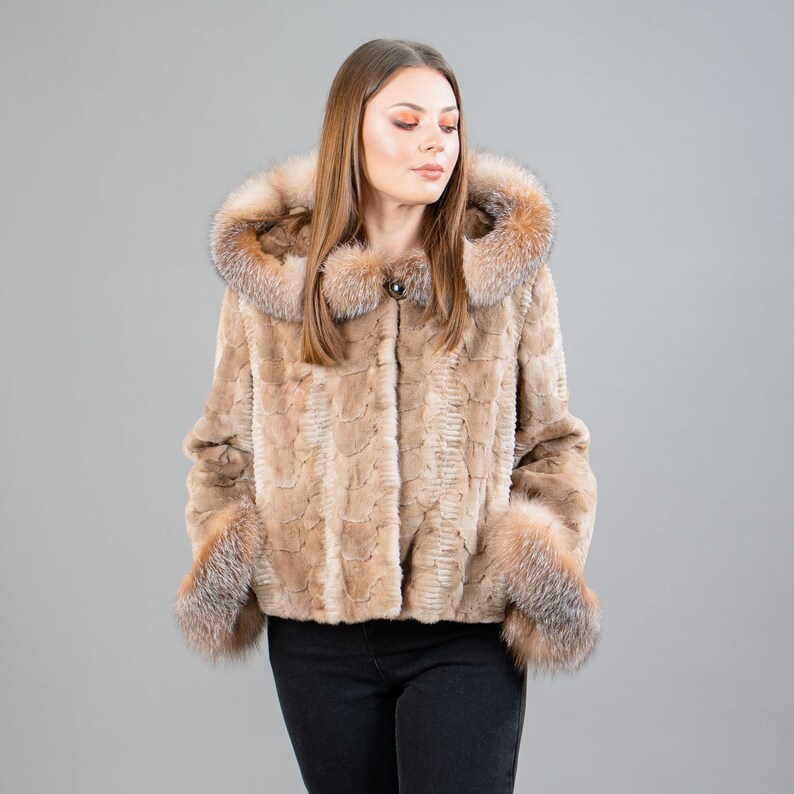 A female model poses in front of a gray panel. She wears a mink fur jacket in beige colors. This short mink fur jacket has a hood, bell-type sleeves, outer pockets, and button closure. The hood and the bell-type sleeves have beige fox fur.