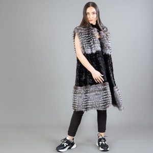 A female model is posing in front of a gray panel. She is wearing a vest made from real fox and mink fur in black and silver colors.