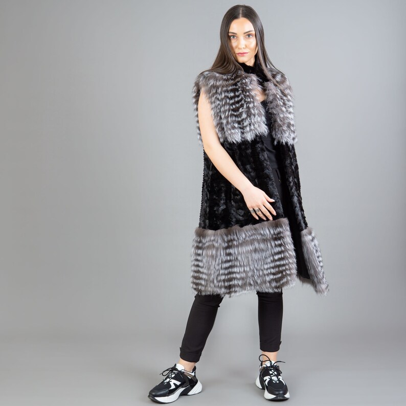 A female model is posing in front of a gray panel. She is wearing a vest made from real fox and mink fur in black and silver colors.