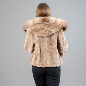 A female model poses in front of a gray panel. She wears a mink fur jacket in beige colors. This short mink fur jacket has a hood, bell-type sleeves, outer pockets, and button closure. The hood and the bell-type sleeves have beige fox fur.