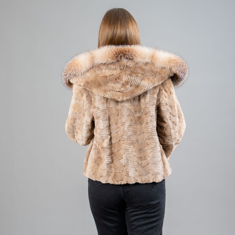 A female model poses in front of a gray panel. She wears a mink fur jacket in beige colors. This short mink fur jacket has a hood, bell-type sleeves, outer pockets, and button closure. The hood and the bell-type sleeves have beige fox fur.