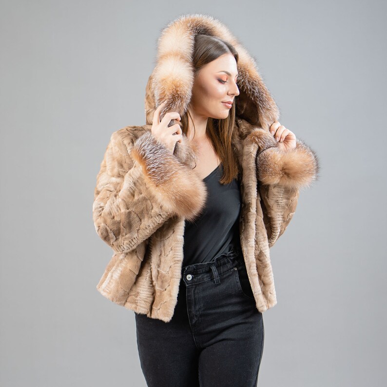 A female model poses in front of a gray panel. She wears a mink fur jacket in beige colors. This short mink fur jacket has a hood, bell-type sleeves, outer pockets, and button closure. The hood and the bell-type sleeves have beige fox fur.