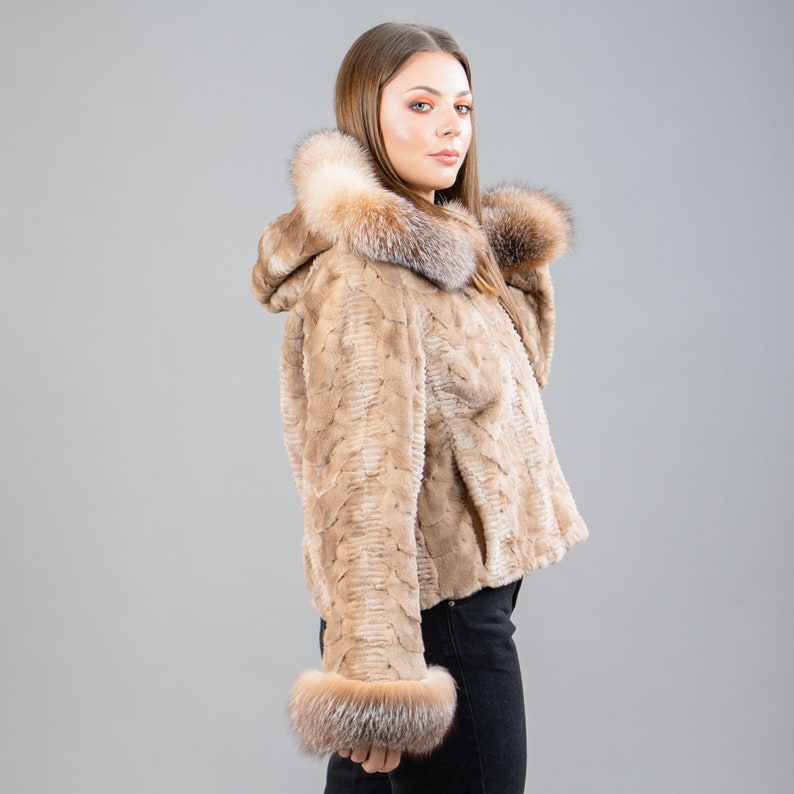 A female model poses in front of a gray panel. She wears a mink fur jacket in beige colors. This short mink fur jacket has a hood, bell-type sleeves, outer pockets, and button closure. The hood and the bell-type sleeves have beige fox fur.