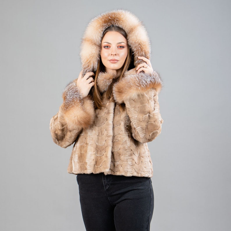 A female model poses in front of a gray panel. She wears a mink fur jacket in beige colors. This short mink fur jacket has a hood, bell-type sleeves, outer pockets, and button closure. The hood and the bell-type sleeves have beige fox fur.