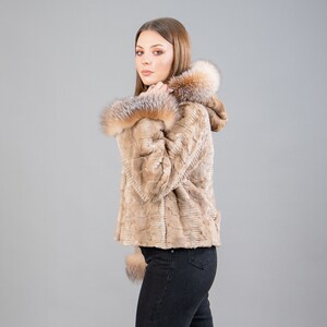 A female model poses in front of a gray panel. She wears a mink fur jacket in beige colors. This short mink fur jacket has a hood, bell-type sleeves, outer pockets, and button closure. The hood and the bell-type sleeves have beige fox fur.