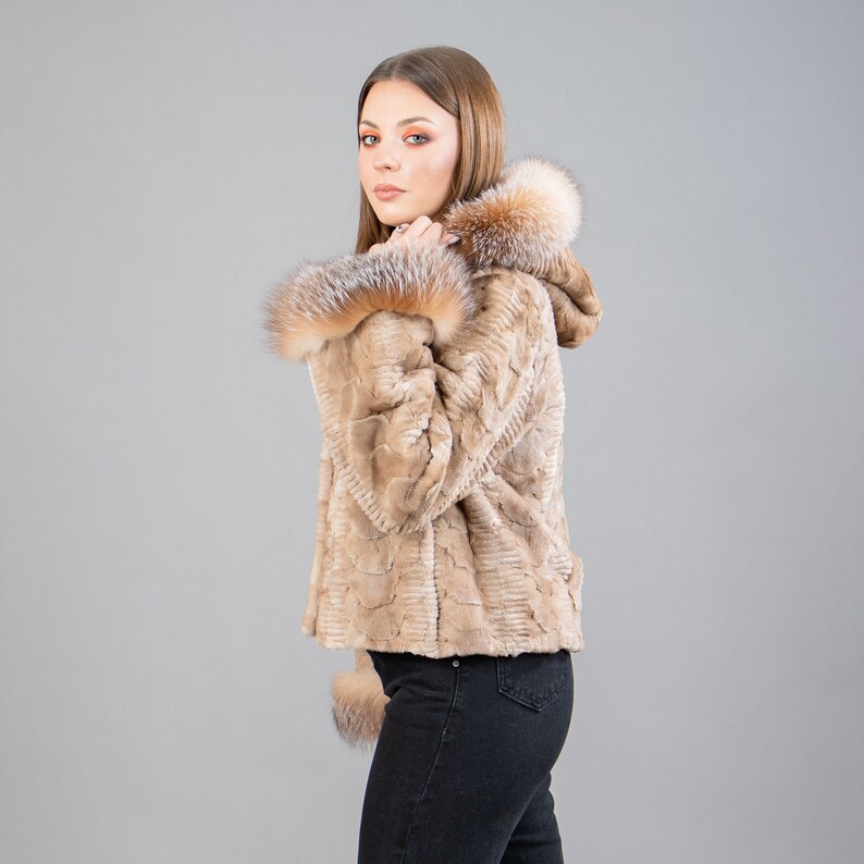 A female model poses in front of a gray panel. She wears a mink fur jacket in beige colors. This short mink fur jacket has a hood, bell-type sleeves, outer pockets, and button closure. The hood and the bell-type sleeves have beige fox fur.