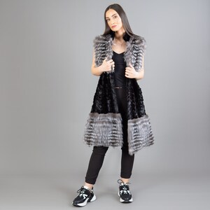 A female model is posing in front of a gray panel. She is wearing a vest made from real fox and mink fur in black and silver colors.