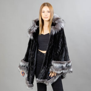 A model posing in front of a gray panel wearing a three-quarters long mink fur coat in black color. The coat has a silver fox fur hood and silver fox fur trim on the bell-type sleeves, and the bottom end.