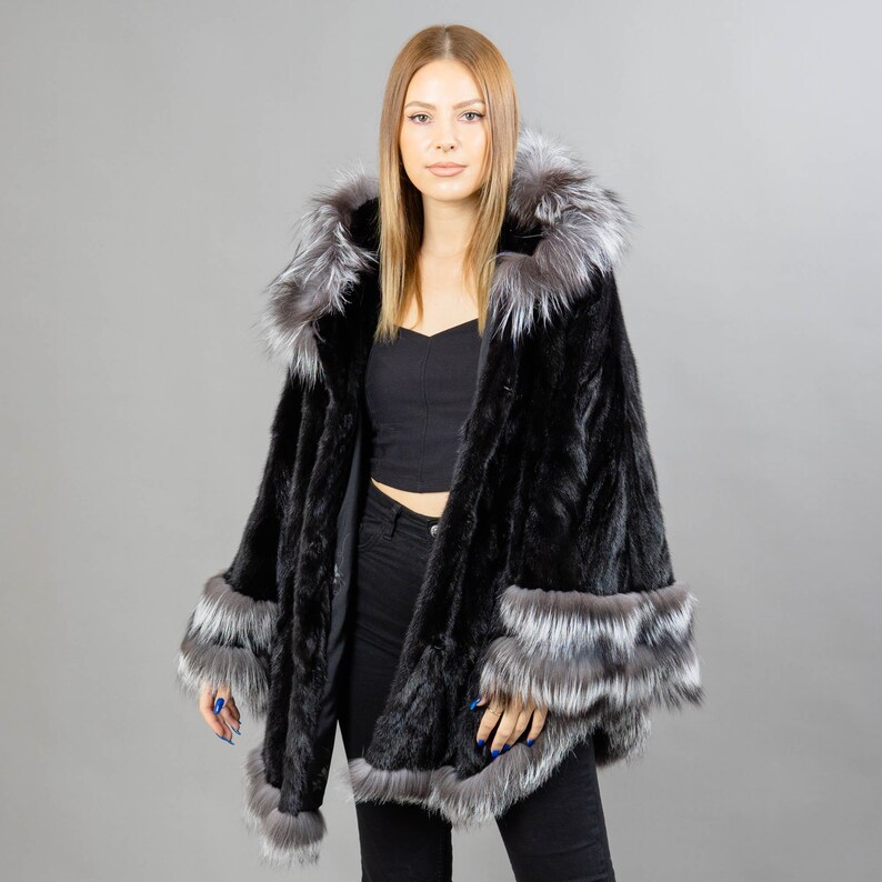 A model posing in front of a gray panel wearing a three-quarters long mink fur coat in black color. The coat has a silver fox fur hood and silver fox fur trim on the bell-type sleeves, and the bottom end.