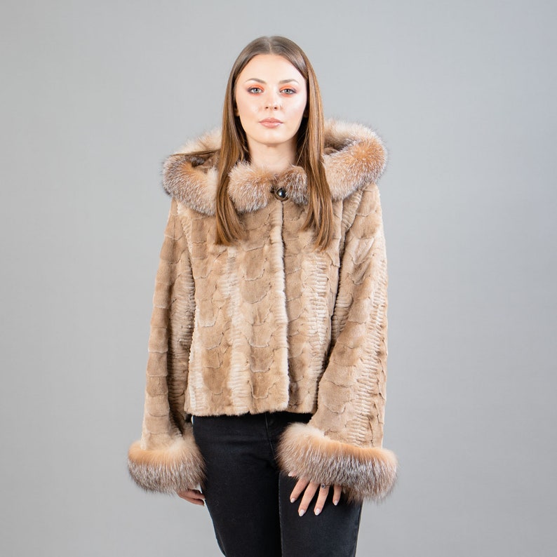 A female model poses in front of a gray panel. She wears a mink fur jacket in beige colors. This short mink fur jacket has a hood, bell-type sleeves, outer pockets, and button closure. The hood and the bell-type sleeves have beige fox fur.