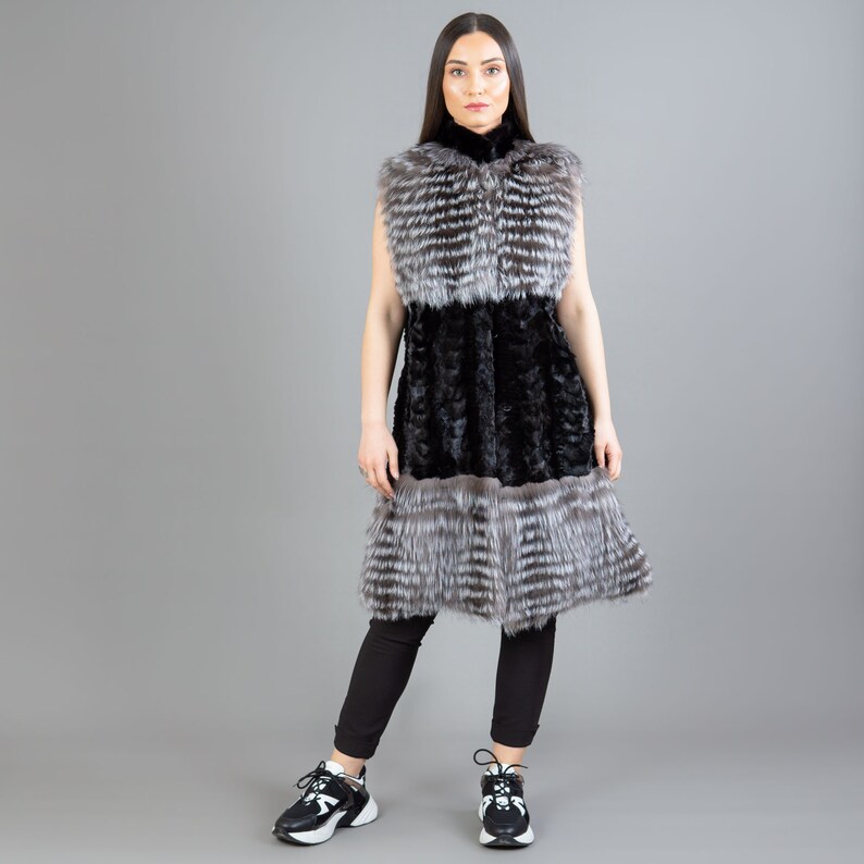 A female model is posing in front of a gray panel. She is wearing a vest made from real fox and mink fur in black and silver colors.