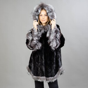 A model posing in front of a gray panel wearing a three-quarters long mink fur coat in black color. The coat has a silver fox fur hood and silver fox fur trim on the bell-type sleeves, and the bottom end.