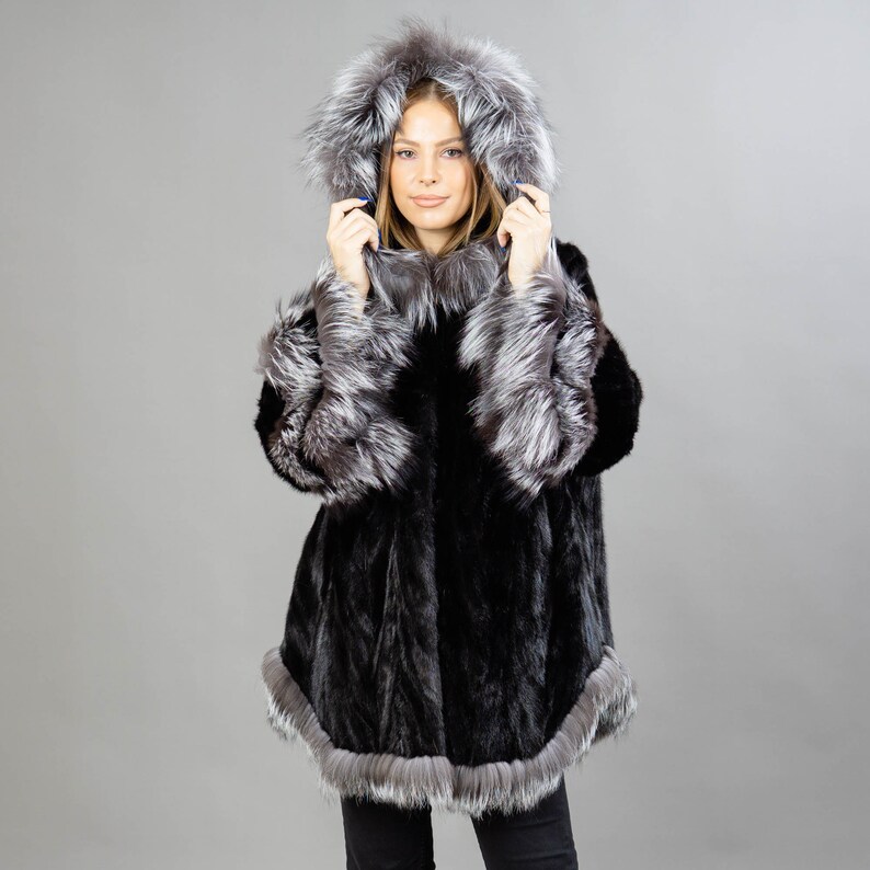 A model posing in front of a gray panel wearing a three-quarters long mink fur coat in black color. The coat has a silver fox fur hood and silver fox fur trim on the bell-type sleeves, and the bottom end.
