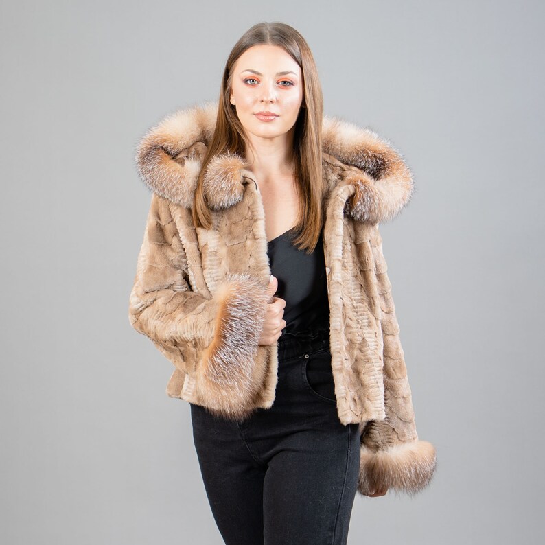 A female model poses in front of a gray panel. She wears a mink fur jacket in beige colors. This short mink fur jacket has a hood, bell-type sleeves, outer pockets, and button closure. The hood and the bell-type sleeves have beige fox fur.