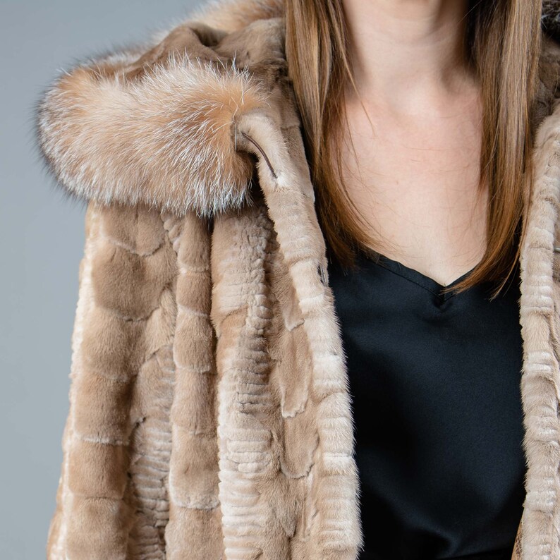 A female model poses in front of a gray panel. She wears a mink fur jacket in beige colors. This short mink fur jacket has a hood, bell-type sleeves, outer pockets, and button closure. The hood and the bell-type sleeves have beige fox fur.