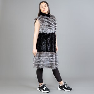 A female model is posing in front of a gray panel. She is wearing a vest made from real fox and mink fur in black and silver colors.