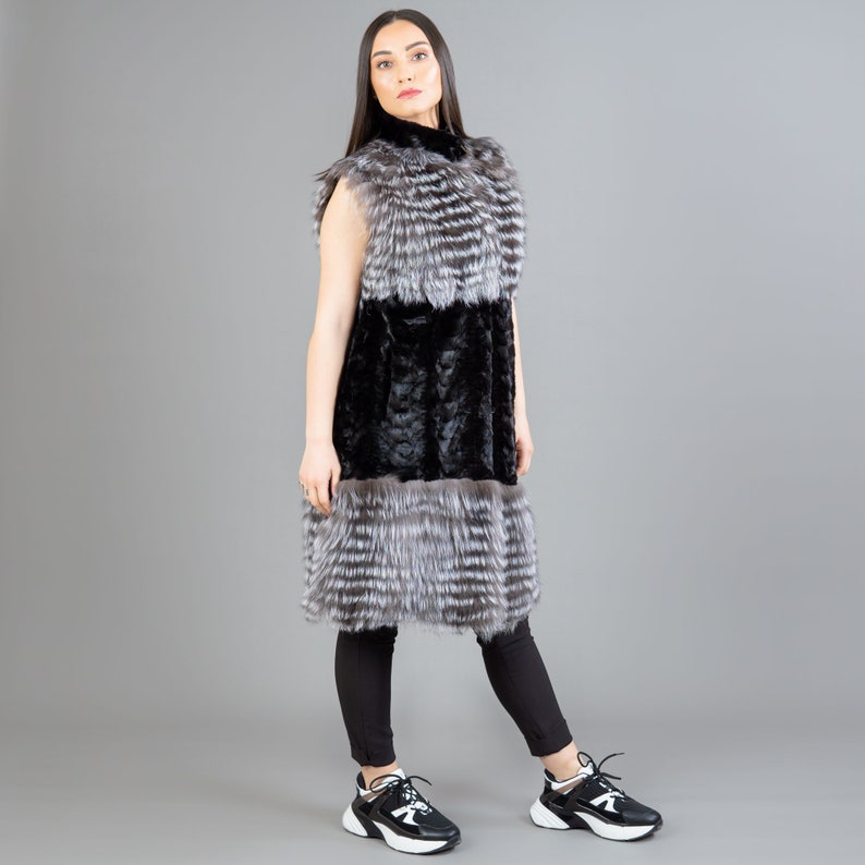 A female model is posing in front of a gray panel. She is wearing a vest made from real fox and mink fur in black and silver colors.