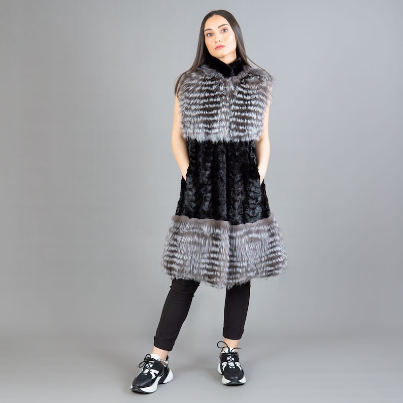 A female model is posing in front of a gray panel. She is wearing a vest made from real fox and mink fur in black and silver colors.