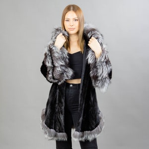 A model posing in front of a gray panel wearing a three-quarters long mink fur coat in black color. The coat has a silver fox fur hood and silver fox fur trim on the bell-type sleeves, and the bottom end.