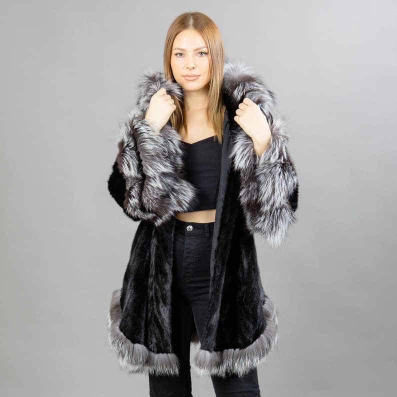 A model posing in front of a gray panel wearing a three-quarters long mink fur coat in black color. The coat has a silver fox fur hood and silver fox fur trim on the bell-type sleeves, and the bottom end.