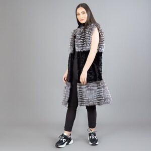A female model is posing in front of a gray panel. She is wearing a vest made from real fox and mink fur in black and silver colors.